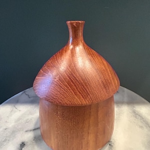 以下が含まれることがあります： 蓋付きのどんぐり型の木製ボックス。ボックスは滑らかな仕上げの明るい茶色の木でできています。