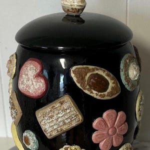 Peut inclure: Pot à biscuits en céramique noire avec couvercle et bouton. Le pot est décoré de diverses formes en céramique, dont un cœur, une fleur et des biscuits. Le pot repose sur une surface en marbre.