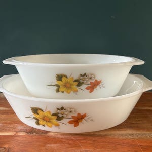 Könnte beinhalten: Zwei weiße Servierschalen mit Griffen, gestapelt auf einer Holzoberfläche. Jede Schale hat ein Blumendesign mit gelben, orangefarbenen und weißen Blumen. Die Schalen haben eine klassische, abgerundete Form, ideal zum Servieren von Gerichten.