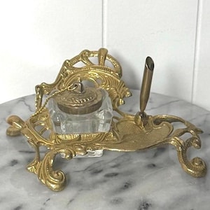 May include: An antique-style inkwell set in gold-tone metal, featuring a clear glass ink pot and a pen holder. The design includes elaborate scrollwork and is presented on a marble surface. The lid is attached to the pot by a small chain.