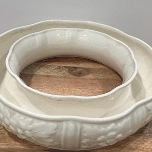 Puede incluir: Cuenco central de cerámica blanquecina con borde ondulado y diseño floral en relieve. El cuenco tiene un anillo interior circular y un anillo exterior más ancho. Para uso decorativo o para servir.