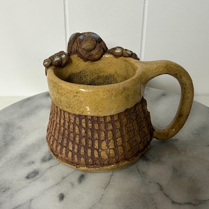 May include: A handmade ceramic mug with a turtle design. The mug has a tan body with a textured brown band around the middle. The turtle is brown with blue accents. The handle is curved and tan.