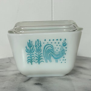 Puede incluir: Cazuela Pyrex blanca con tapa de cristal transparente. El plato presenta un diseño de gallo y flores turquesas. El plato cuadrado mide aproximadamente 15 cm de ancho y 8 cm de alto.
