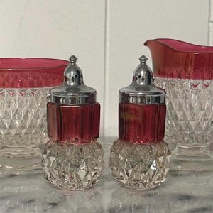 May include: A vintage ruby red and clear glass serving set. The set includes a sugar bowl, a creamer, and two salt and pepper shakers. The glass pieces have a diamond pattern. The sugar bowl and creamer have ruby red rims.