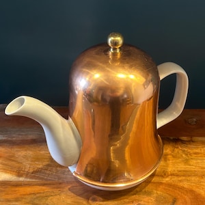 Puede incluir: Una tetera blanca con una tapa de cobre y un pomo de latón. La tetera tiene un cuerpo redondeado y un pico en el lateral. La tapa de cobre es brillante y refleja la luz.