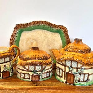 Puede incluir: Un encantador juego de té de cerámica con tetera, azucarero, jarra de crema y una bandeja. Las piezas tienen forma de cabañas pintorescas con tejados de paja, puertas marrones y detalles de ventanas. La bandeja tiene un borde verde y marrón.