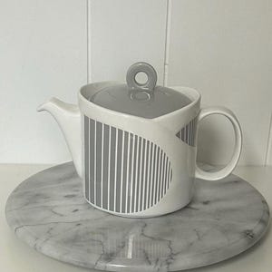Può includere: Una teiera in ceramica bianca con coperchio e manico grigi. La teiera presenta un motivo a righe grigie e bianche sul lato. La teiera è appoggiata su un vassoio rotondo di marmo.