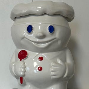 Pode incluir: Um pote de biscoitos de cerâmica branca em forma de um chef sorridente com olhos azuis e uma colher vermelha. O chef usa um chapéu e uniforme brancos com botões vermelhos. Um item de decoração de cozinha vintage.