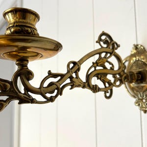 Könnte beinhalten: Verzierte Wandleuchte aus Messing mit Kerzenhalter und dekorativen Schnörkeln. Die Leuchte hat einen kreisförmigen Sockel und einen geschwungenen Arm mit floralen und Blattmotiven. Das Messing hat eine warme, gealterte Patina.
