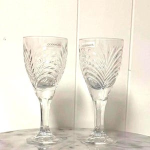 May include: Two clear crystal wine glasses with etched leaf patterns. The glasses have long stems and round bases. The brand name "GODINGER" is visible on the upper part of the glasses. The glasses are on a marble surface.
