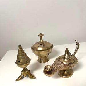 Op de afbeelding: Drie antieke messing objecten: een kegelvormige wierookbrander, een ronde wierookbrander met een deksel en een genielamp met geëtste details. De items worden op een wit oppervlak weergegeven.