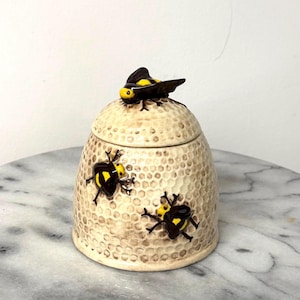 May include: A ceramic honey pot with a lid, featuring a textured honeycomb pattern and three-dimensional bee embellishments. The bees are black and yellow. The pot is beige and sits on a marble surface.