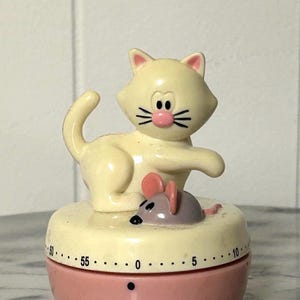 May include: A vintage kitchen timer shaped like a cream-colored cat with a pink nose and a gray mouse on top. The timer has a pink base with a white dial and black numbers, measuring up to 60 minutes.