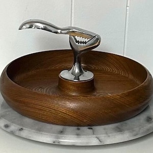 May include: A wooden bowl with a stainless steel nutcracker and a marble base. The bowl is a rich brown colour with a circular grain pattern. The nutcracker is silver and has a claw-like design.