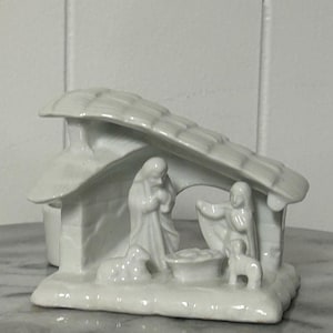 Puede incluir: Un belén de cerámica blanca que representa a María, José y el niño Jesús en un pesebre, con una estructura techada. También hay un cordero y un perro. Las figuras son detalladas y la escena está sobre mármol.