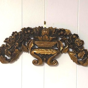 May include: An ornate, gold-coloured wall decoration featuring a central urn design flanked by floral and leaf carvings. The decorative piece is mounted on a white surface, showcasing its detailed craftsmanship.