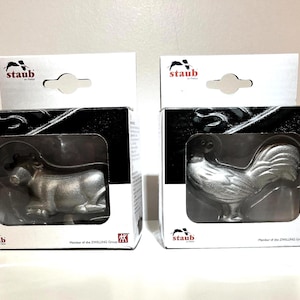 Puede incluir: Dos figuras de Staub en cajas. Una es un toro plateado, la otra un gallo. Las cajas son negras con tapas blancas y el logotipo de Staub. Las figuras son visibles a través de plástico transparente.