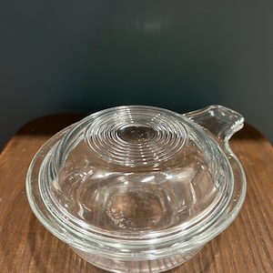 Puede incluir: Un plato para hornear de vidrio transparente con tapa. El plato es redondo y tiene un asa en el lateral. La tapa tiene una serie de círculos concéntricos en la parte superior.