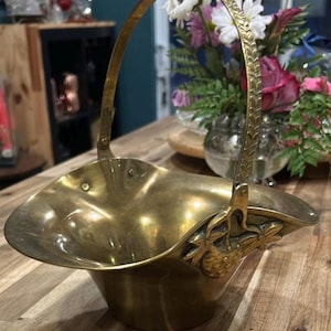 Könnte beinhalten: Ein Vintage-Messingkorb mit gebogenem Griff und floralen Verzierungen. Der Korb hat eine breite, flache Schalenform und steht auf einer Holzoberfläche. Das Messing hat einen warmen, goldenen Farbton.