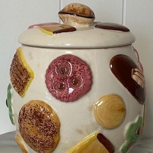Peut inclure: Un bocal à biscuits en céramique vintage avec un couvercle, décoré de motifs en relief colorés de diverses pâtisseries. Le bocal est blanc cassé avec une variété de formes et de couleurs, dont le marron, le jaune, le rose et le vert. Le couvercle a une petite poignée en forme de tarte, marron.