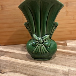 Könnte beinhalten: Eine grüne Keramikvase mit fächerförmigem Oberteil und Schleifendetail. Die Vase hat einen abgerundeten Boden und zwei kleine Griffe. Die Vase ist etwa 20 cm hoch.