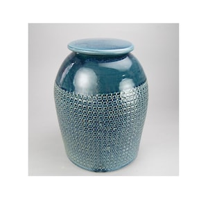 May include: A teal ceramic jar with a lid. The jar has a textured, patterned lower half and a smooth, glossy upper half. The lid is a lighter shade of teal. The jar is a decorative item.