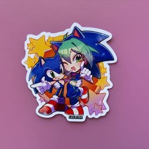 May include: A sticker featuring two cartoon characters, one resembling Sonic the Hedgehog, the other with green hair. They are hugging and pointing, surrounded by yellow stars. The sticker has a white border and the text "SAEWIM".