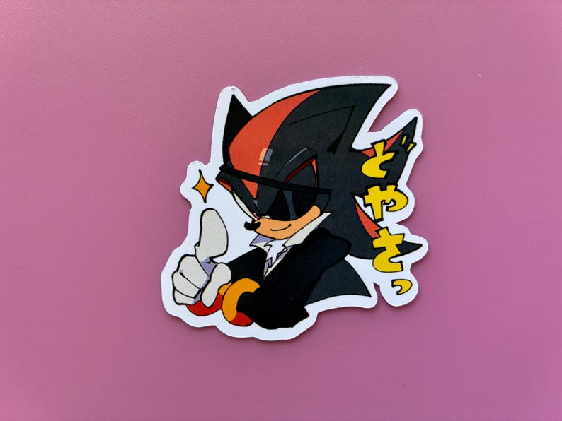 May include: A sticker featuring a cartoon character with black and red spiky hair, wearing sunglasses and a black suit. The character gives a thumbs-up gesture, with Japanese text to the right. A yellow star is next to the character.