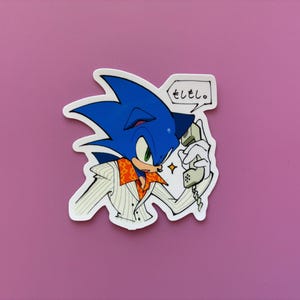May include: A sticker featuring Sonic the Hedgehog in a white jacket with an orange collar, holding a phone. The blue hedgehog has a speech bubble with Japanese text. The sticker is on a pink background.