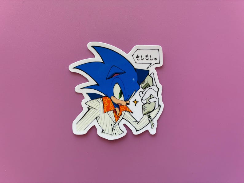 May include: A sticker featuring Sonic the Hedgehog in a white jacket with an orange collar, holding a phone. The blue hedgehog has a speech bubble with Japanese text. The sticker is on a pink background.