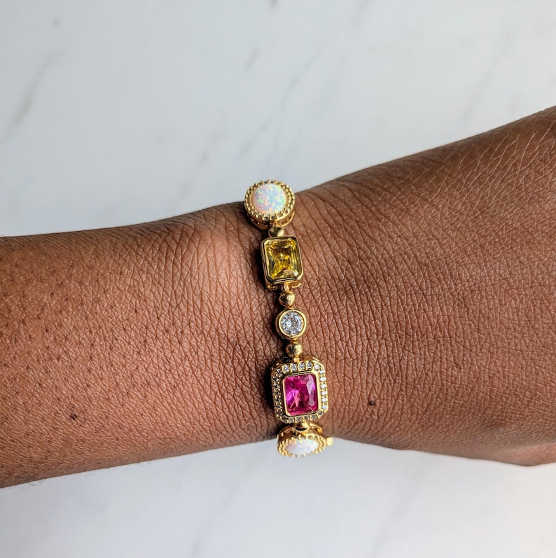 May include: Gold-toned bracelet featuring a series of colorful gemstones. The bracelet includes a round opal, a square yellow stone, a small clear stone, a square pink stone, and a second round opal. The bracelet is worn on a wrist.