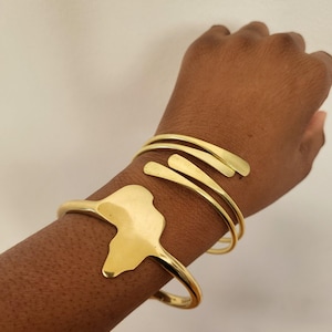 May include: Gold-toned metal bracelets. One bracelet features the outline of the African continent. The other bracelet has a spiral design with three parallel bars. The bracelets are worn on the wrist.