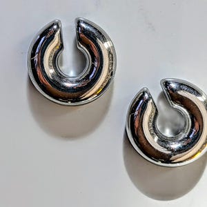 May include: Two polished silver ear weights, each with a curved, horseshoe shape and a split at the top. The weights reflect light, showing a mirrored surface. They are presented on a white background. Each weight is approximately 2.5 cm in diameter.