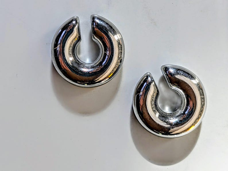 May include: Two polished silver ear weights, each with a curved, horseshoe shape and a split at the top. The weights reflect light, showing a mirrored surface. They are presented on a white background. Each weight is approximately 2.5 cm in diameter.