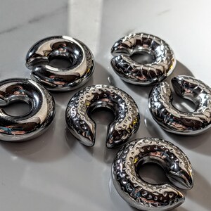 May include: Six shiny, silver-coloured, horseshoe-shaped metal objects are arranged on a light-coloured surface. The objects have a smooth, polished appearance, with a circular design and a split opening. Some have a textured surface.