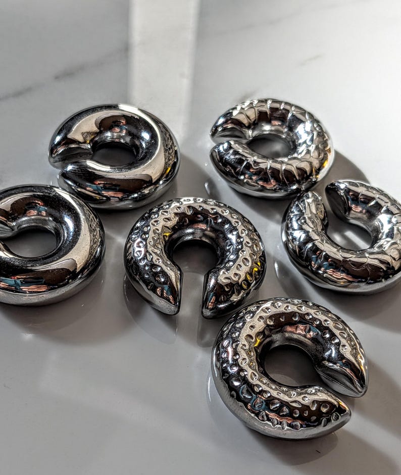 May include: Six shiny, silver-coloured, horseshoe-shaped metal objects are arranged on a light-coloured surface. The objects have a smooth, polished appearance, with a circular design and a split opening. Some have a textured surface.