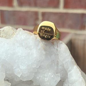 Puede incluir: Un anillo de sello de oro con una cara cuadrada que dice "made of magic". El anillo está sobre un cristal blanco.