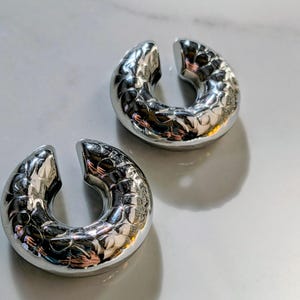May include: Two silver-coloured, U-shaped ear cuffs with a textured, patterned surface. The cuffs are open at one end and sit on a white surface. The jewellery reflects light, creating highlights and shadows.