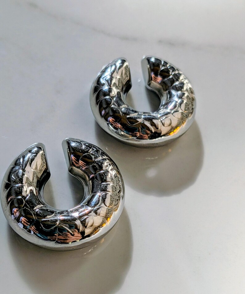 May include: Two silver-coloured, U-shaped ear cuffs with a textured, patterned surface. The cuffs are open at one end and sit on a white surface. The jewellery reflects light, creating highlights and shadows.