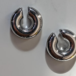 May include: Two polished, silver ear weights. Each weight has a rounded, horseshoe-like shape with a split at the top. The weights are reflective, with the surrounding environment mirrored on their surfaces.