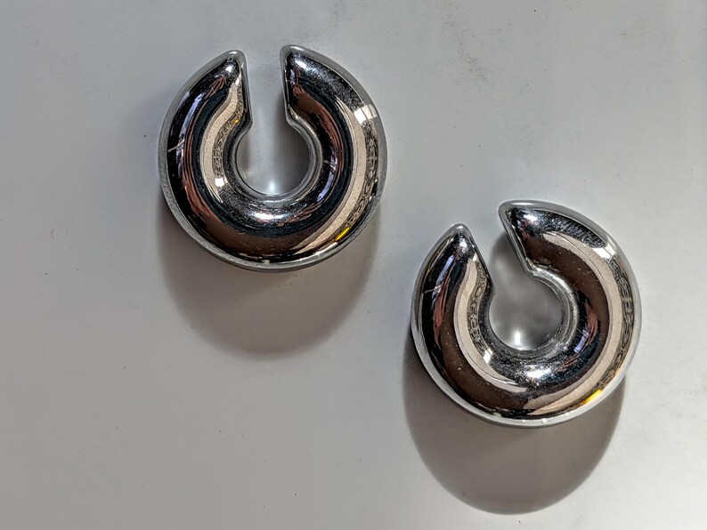 May include: Two polished, silver ear weights. Each weight has a rounded, horseshoe-like shape with a split at the top. The weights are reflective, with the surrounding environment mirrored on their surfaces.
