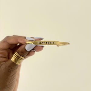 May include: A gold-colored bracelet with the words "STAY SOFT" engraved on it. The bracelet has a simple, elegant design and is held up against a neutral background. Several gold rings are visible on the hand.