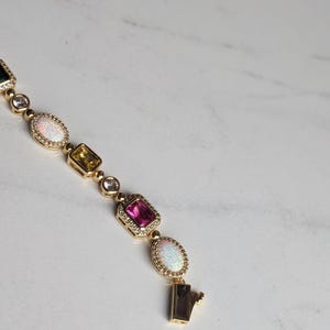 May include: Gold bracelet featuring a series of colorful gemstones, including pink, yellow, and white opalescent stones. The bracelet has a secure clasp and is set against a white marble background.