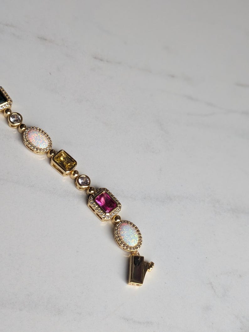 May include: Gold bracelet featuring a series of colorful gemstones, including pink, yellow, and white opalescent stones. The bracelet has a secure clasp and is set against a white marble background.
