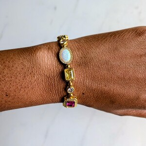 May include: Gold-toned bracelet featuring an oval opal, a yellow square stone, a clear round stone, and a pink square stone. The bracelet is adorned with small, clear accent stones and is worn on a brown skin tone.