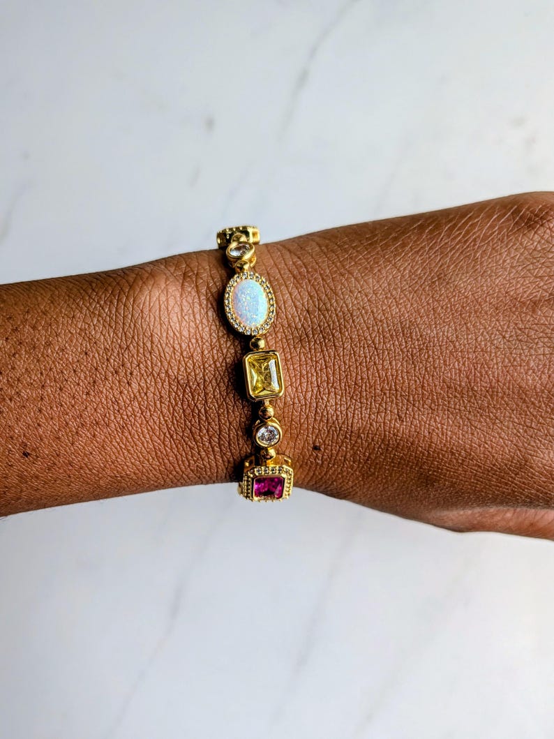 May include: Gold-toned bracelet featuring an oval opal, a yellow square stone, a clear round stone, and a pink square stone. The bracelet is adorned with small, clear accent stones and is worn on a brown skin tone.