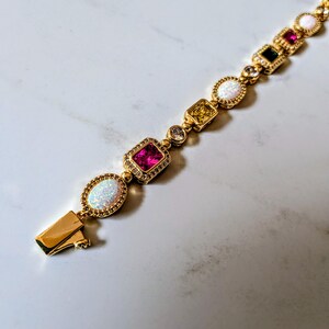 May include: Gold bracelet featuring a variety of colorful gemstones, including pink, white, yellow, and black stones. The bracelet has a secure clasp and is set against a white marble background. The bracelet is a piece of jewelry.