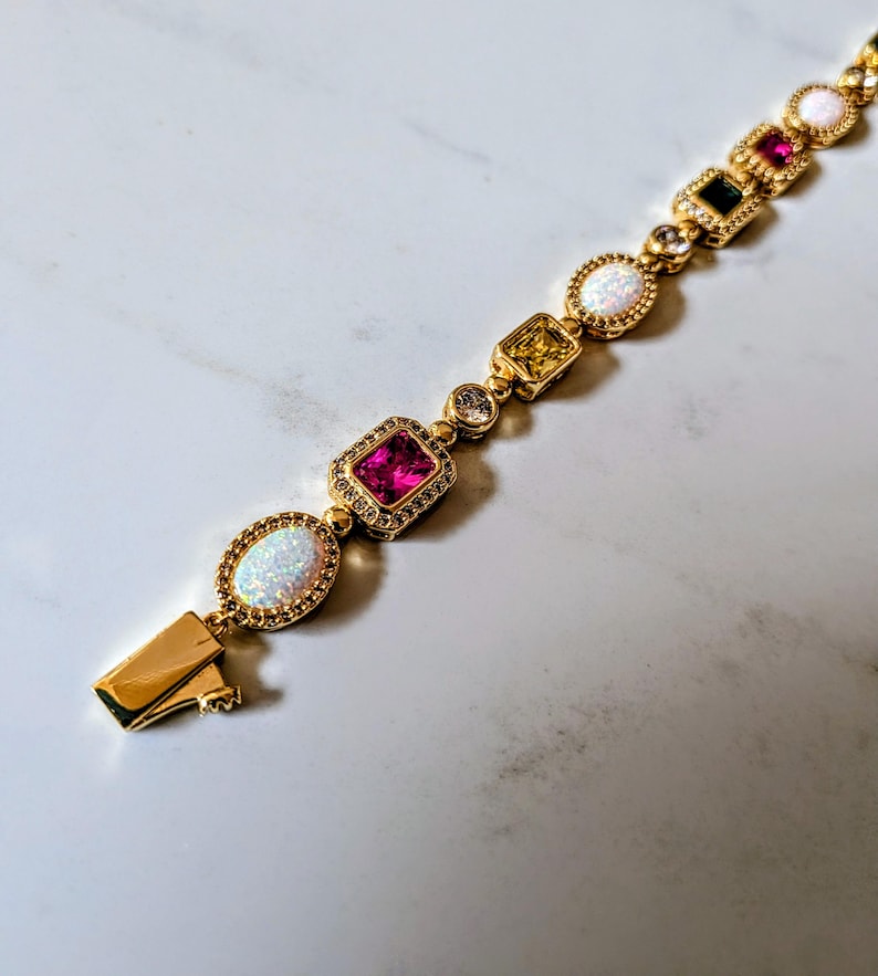 May include: Gold bracelet featuring a variety of colorful gemstones, including pink, white, yellow, and black stones. The bracelet has a secure clasp and is set against a white marble background. The bracelet is a piece of jewelry.
