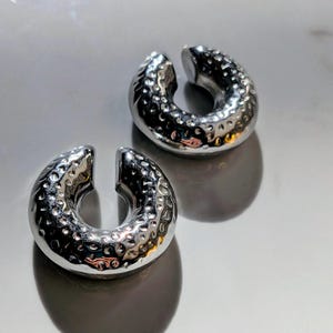 May include: Two silver ear cuffs with a hammered metal texture. The C-shaped cuffs are open-ended and sit on a reflective surface. The polished finish of the cuffs reflects light, highlighting their design and texture.