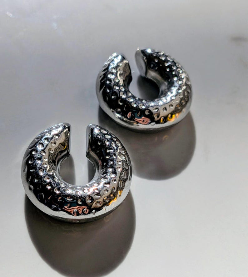 May include: Two silver ear cuffs with a hammered metal texture. The C-shaped cuffs are open-ended and sit on a reflective surface. The polished finish of the cuffs reflects light, highlighting their design and texture.
